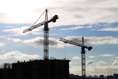İnşaat vinçlerinin siluetleri ve bulutlu mavi gökyüzüne karşı işçiler. Konut inşaatı, şehirdeki apartman bloğu