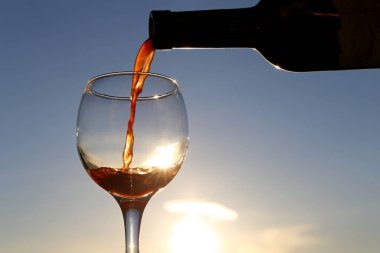 Kırmızı şarap, güzel günbatımı arka planında bardağa dökülüyor. Güneş şarap kadehinde parlıyor. Kutlama konsepti, tatil köyündeki yaz partisi, dışarıda romantik bir akşam yemeği.