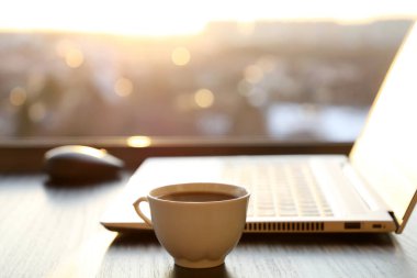 İş yerindeki sıcak kahve fincanı ve dizüstü bilgisayarla gün batımında pencerenin önünde. Uzaktan çalışma ve çevrimiçi kazanç kavramı