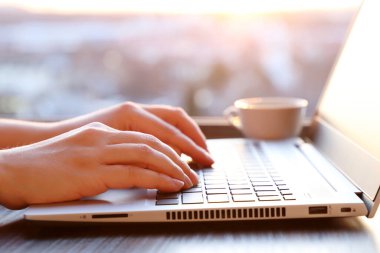 Gün batımında pencerenin yanındaki dizüstü bilgisayarın başında oturan kadın, kadın elleri güneş ışığında yakınlaşıyor. Kahve fincanı, internet iletişimi, ofis veya ev işi konsepti olan rahat bir iş yeri