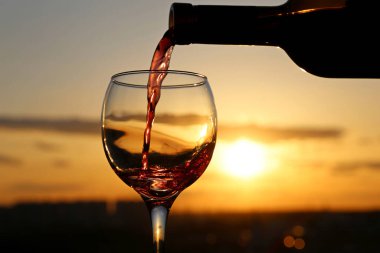 Kırmızı şarap güzel bir günbatımı arka planında bardağa dökülüyor. Kutlama konsepti, tatil köyündeki yaz partisi, dışarıda romantik bir akşam yemeği.