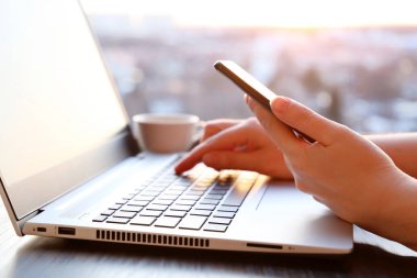 Akıllı telefonlu bir kadın günbatımında pencerenin yanında dizüstü bilgisayarın başında oturuyor. Konforlu iş yeri, çevrimiçi iletişim, ofis veya ev işi ve ödeme kavramı