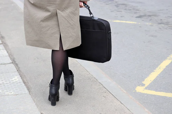 Female legs stockings and boots on high heels on the street. Elegant woman in coat standing with a briefcase on bus stop in spring