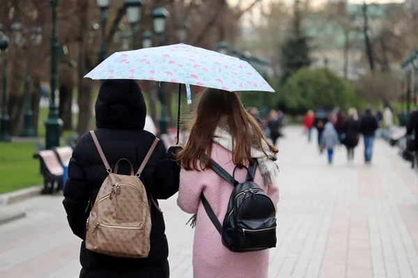 Rain in city, two girls with one umbrella walk on a street on people background. Rainy weather, spring storm
