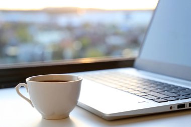 İş yerindeki sıcak kahve fincanı ve dizüstü bilgisayarla gün doğumunda pencerenin önünde. Günaydın, uzaktan çalışma ve online kazanç kavramı.