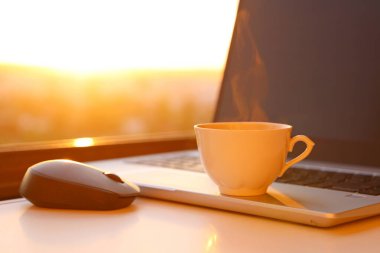 Gün batımında sıcak kahve fincanı ve dizüstü bilgisayar pencerenin önünde. Ev ofisindeki rahat iş yeri, uzaktan çalışma ve online kazanç kavramı