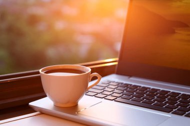 Günbatımında dizüstü bilgisayarın üzerinde kahve fincanı. Ev ofisindeki rahat iş yeri, uzaktan çalışma kavramı.