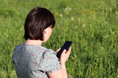 Kablosuz kulaklıklı bir kadın yaz otlağında akıllı telefon kullanıyor. Yeşil arka planda cep telefonu olan kadın eller