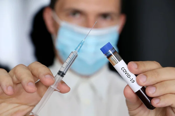 Doctor in mask with syringe and covid-19 blood sample in test tube in hands. Concept of vaccination and medical testing