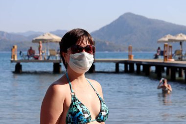 Woman in protective mask and sunglasses standing on a sea beach on pier and mountains background. Safety at tourist resort during coronavirus pandemic