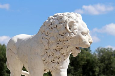 Taş aslan yaz parkını dekore ediyor. Mavi gökyüzünde mermer heykel ve arka planda yeşil ağaçlar