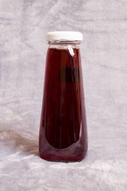 Kırmızı meyve suyu cam şişede, gri arka planda.