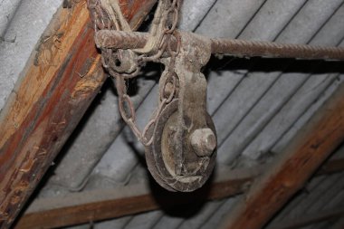 Metal çatılı eski bir yapının içindeki ahşap kirişlerden paslı bir makara asılıdır. Ayar yaş ve yıpranma belirtileri gösteriyor. Havada toz görünüyor..