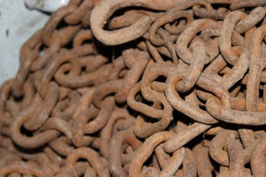 Birçok paslı metal zincir bir depo alanında istiflenir. Zincirler birbirine dolanmış ve düzensiz, yüzeyi biraz toprak ve toz kaplamış. Sahne aydınlık görünüyor..