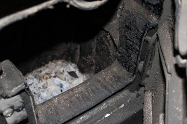 Bir odun sobasının içinde kül ve is kalıntıları var. Yüzeyde karanlık maddeler vardır ve metal parçalarıyla çevrilidir. Sahne gölgelerle kararmış..