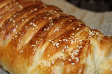 Üzerinde altın kahverengi kabuk ve susam tohumları olan uzun bir hamur işi parşömen kağıdının üzerinde duruyor. Sıcak ışıklandırması olan bir mutfakta tezgahın üstüne konur..