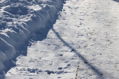 Temiz bir yol, büyük bir kar yığınının yanında taze kar üzerinde ayak izleri gösteriyor. Güneş ışığı karların üzerine gölge düşürür. Bu sahne mahallede tipik bir kıştır..