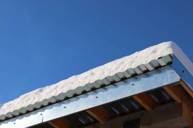 Kar, ahşap kirişli bir binanın metal çatısında duruyor. Gökyüzü açık ve mavi, bulutsuz soğuk bir kış gününü gösteriyor..