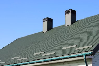 Bir çatının açık mavi bir gökyüzünün altında iki bacası vardır. Çatıda metal paneller ile yeşil bir renk var. Görüntü basit bir ev yapısını ve açık havayı gösteriyor..