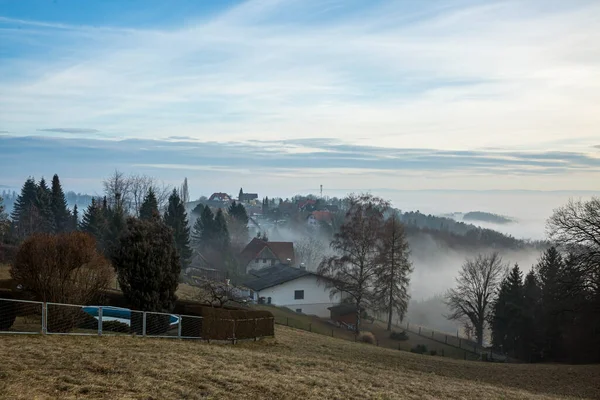 Malerischer Blick auf das grüne Tal mit kleinem Dorf umgeben von Bäumen in der Nähe der Bergkette der Alpen in nebligen Morgen. — Stockfoto