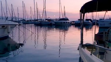 Rıhtıma yanaşmış yelkenli tekneleri ve yatları olan hareketli bir marinanın üzerinde sakin bir günbatımı. Pembe ve mor gökyüzü sakin suya hafifçe yansıyarak huzurlu bir deniz manzarası yaratıyor..