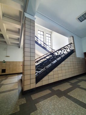 Geniş merdiveni, süslü metal parmaklıkları, geniş çok camlı pencereleri ve desenli döşemeleri olan eski bir döşeme. Parlak doğal ışık, mimari detaylar ve zamansız kamu binası atmosferi.