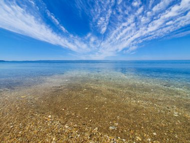 Parlak mavi gökyüzünün altında çakıl taşları ve kristal suyla temiz, sığ sahil şeridi ve ince bulutlar, sakin yaz rahatlığı, seyahat, dinginlik ve doğal kıyı güzelliğini çağrıştırıyor..