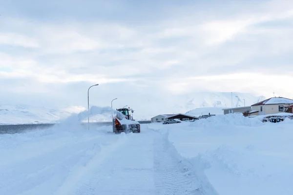 The tractor cleans snow on the road after a heavy snowfall. A snow blower removes snow from the road in the morning. Winter landscape of a small town in Iceland.