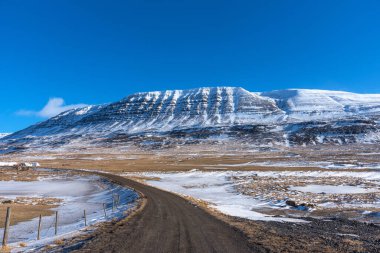 Tipik İzlanda kış manzarası. Kar altında dağlar ve mavi gökyüzü. Dağlarla kaplı güzel kış manzarası, soğuk soğuk hava ve İzlanda 'da beyaz bir tarlada asfalt yol..