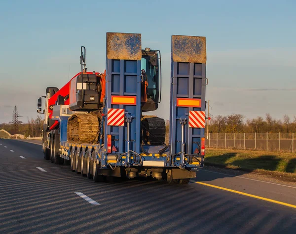 truck transports an excavator on a trailer along a suburban highway