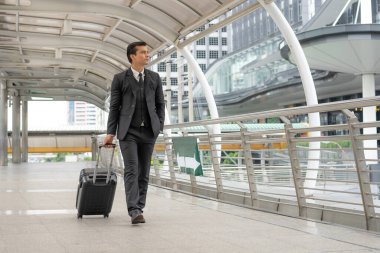 Siyah takım elbiseli genç bir iş adamı köprüde bir bavulu sürüklüyor, modern binaya bakıyor, mekanı kopyalıyor.