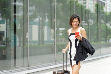 Genç, güzel Asyalı bir kadın akıllı telefon kullanıyor ve yürüyüş yolunda bavuluyla mutlu bir şekilde yürüyor.