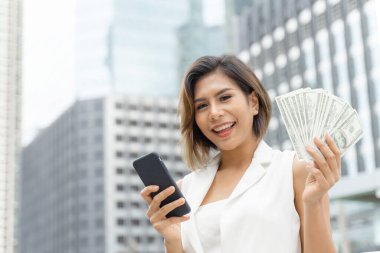 Başarılı bir Asyalı iş kadını akıllı telefon ve para kullanıyor. Amerikan dolarları, iş konsepti.