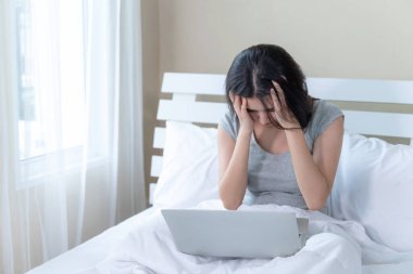 Yatakta oturan genç ve güzel bir kadın dizüstü bilgisayar kullanıyor. Ciddi ve baş ağrısı çekiyor.