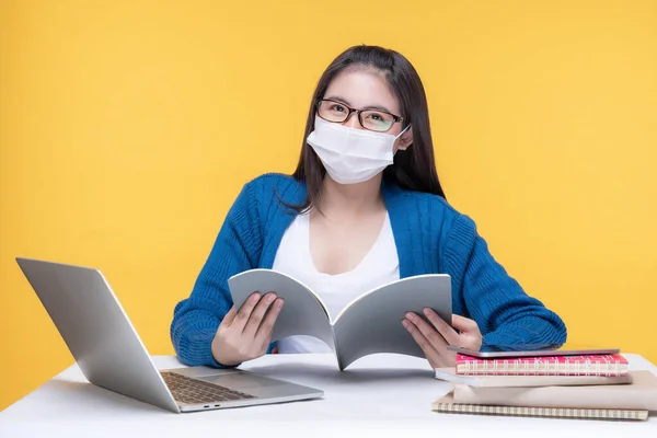 Portrait of a beautiful young woman student with glasses wear a mask holding textbook on yellowbackground - studying online e-learning system during the corona virus covid19 outbreak
