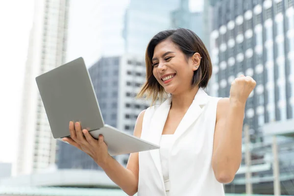Young pretty working woman with action winner and happily with successful on laptop in hand, Blurred background with modern building in city - business concept
