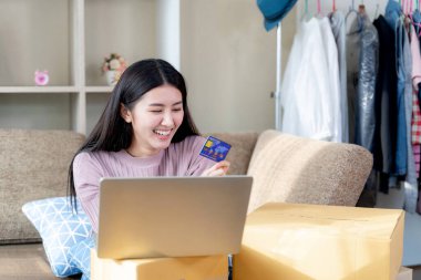 Evdeki oturma odasındaki kanepede oturan genç ve mutlu bir kadın. Gülümsüyor ve kredi kartıyla internetten alışveriş yapıyor. Arka planda bulanık askı bezleri var.