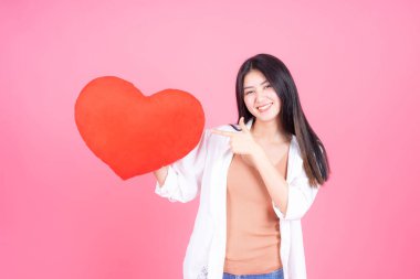 Başarılı bir Asyalı genç kadın pembe arka planda kırmızı kalp şeklinde yastığı elinde tutuyor.
