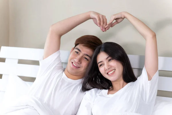 肖像幸せな若いカップルが座ってベッドの上で笑顔と腕を上げる一緒にハートの形を作る — ストック写真