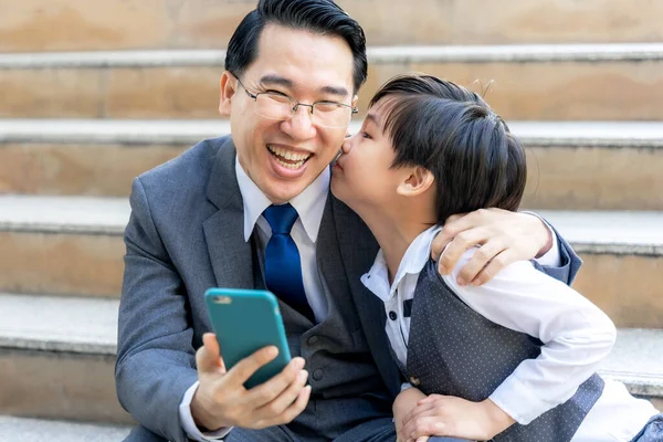 スーツを着たビジネスマンだ 彼の素敵な息子は彼の頬にキスをしていた間 彼は手に携帯電話を見て 喜んで笑った お父さんと息子幸せなアジアの家族の概念 — ストック写真