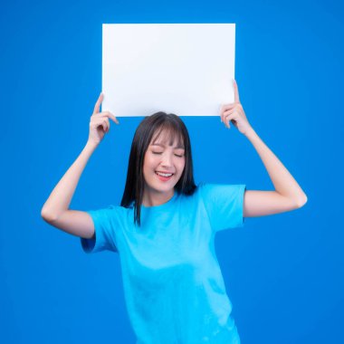 Mavi tişörtlü, kaküllü, güzel Asyalı genç bir kadın. Gülümsüyor ve reklam afişi için boş bir tahta tutuyor. Mavi arka planda izole edilmiş boş bir pano.