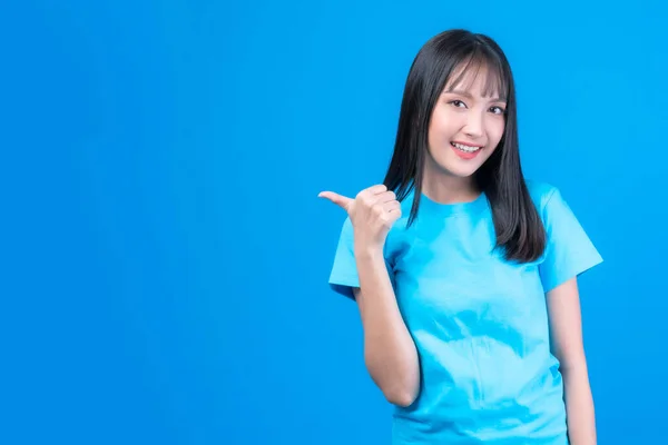 美しいアジアの女性かわいいです女の子とともに前髪のある髪スタイルで青Tシャツ笑顔と指差し指へ空コピースペースのために現在の製品または空白のスペースのために広告のために隔離された青の背景 — ストック写真