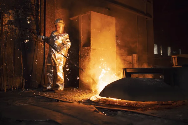 Foundry worker in aluminized protective fire suit checking temperature of molten iron in furnace. Industrial steel production and metallurgy.