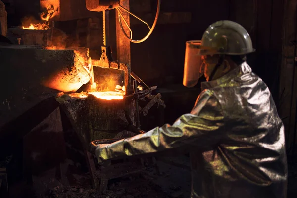 Founder in protective suit working with hot liquid steel in foundry.