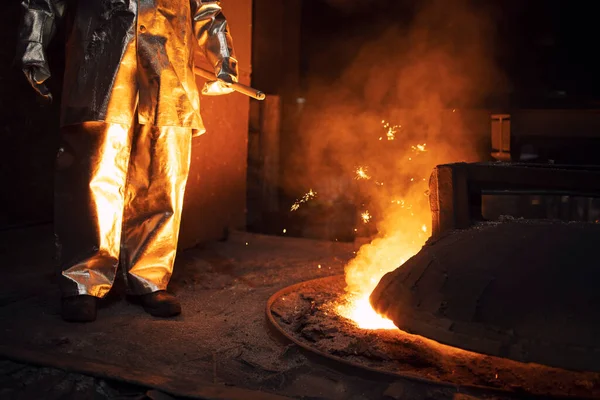 Worker in protection suit controlling iron smelting in furnace and applying heat to ore to extract a base metal. Metallurgy and heavy industry.