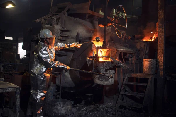 Workman in fire protection suit and hardhat controlling process of melting iron in foundry furnace.