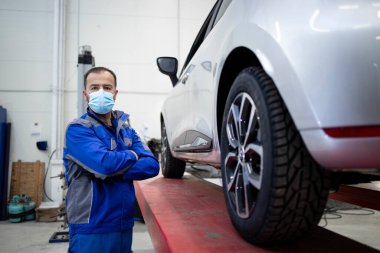 Corona virüsü yüzünden maske takan profesyonel araba tamircisinin portresi bir otomobil atölyesinde duruyor..