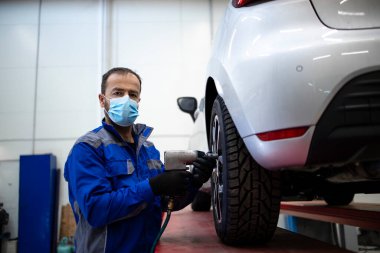 Corona virüsü yüzünden yüz maskesi takan profesyonel araba tamircisinin portresi. Elinde pnömatik vida tabancası ve araç değiştirme tekerleği var. Otomobil servisi ve bakımı.