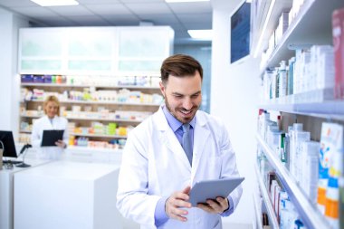 Eczanede ya da eczanede çalışan beyaz önlüklü yakışıklı bir erkek eczacı. Tablet bilgisayarındaki ilaçları kontrol ediyor. Sağlık ve eczacı.