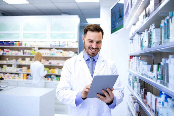 Professional handsome male pharmacist working in pharmacy store or drugstore. Checking medicines on his tablet computer. Healthcare and apothecary.